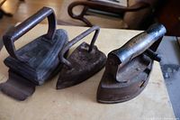 Photo showing all three antique sad irons on a wooden surface, highlighting their aged condition and distinct handles.