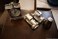 Silver plated clock on left with visible round white clock face with Roman numerals, two detailed vintage car shaped coin banks in center, one salt and one pepper shaker with yin yang style design on right, all displayed on brown surface