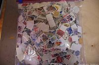 Large transparent plastic bag filled with numerous used postage stamps in various colors and sizes, visibly showing different designs and postmark cancellations.