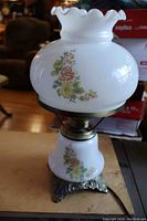 Full lamp view showing floral decorated white glass ruffled top shade, middle metal fixture, and matching floral decorated glass on the base with ornate metal bottom stand.