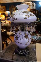 Older colonial style crystal lamp shown unlit, highlighting white milk glass with gold leaf and vine decoration and hanging crystal prisms.