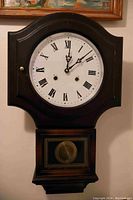 Front view of regulator style wall clock showing white clock face with black Roman numerals and black hands inside a dark wooden arched case.