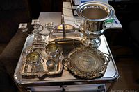 Full set of nine silver plated items arranged on a metal table including candle stand, ice bucket, trays, cream and sugar set, salt cellars, and bread caddy.