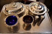 Silver plated serving lot with two lidded dishes, two glass-lined bowls (cobalt blue and ruby red), and salt and pepper shakers with cobalt blue liners on metal table surface.