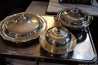 Three silver plate serving pieces with lids shown on a reflective surface. The largest has a glass liner, the middle piece has an enamel liner, and the smallest piece has no liner.