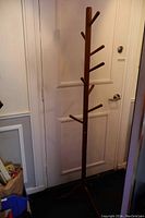 Full view of tall teak wood coat rack against a door, showing vertical pole and wooden pegs for hanging.