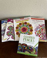 Four adult coloring books arranged on a wooden surface with the set of 50 colored pencils in front. Covers show detailed floral and mandala designs and colorful pencil box.