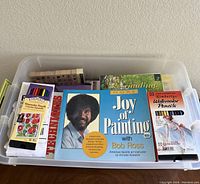 Plastic bin containing art books and art supplies including watercolor pencils and Bob Ross painting books.