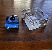 Two glass puzzleweights placed on wooden surface; one larger clear square with label and a smaller blue and black swirl piece.