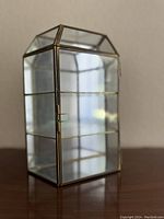 Front diagonal view of vintage brass and glass display case closed, showing its geometric top and glass shelves inside.