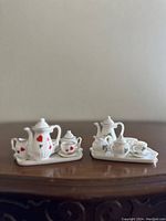 Two miniature porcelain tea sets displayed on a wooden surface, showing full sets including teapot, sugar bowl, creamer, cups, and trays.