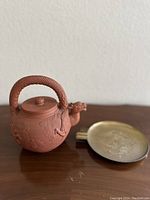 Side angle showing the dragon handle and body details of the clay teapot and the brass dish beside it