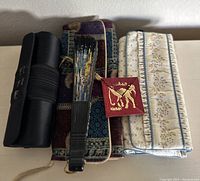 Shows fabric clutches stacked with a black cylindrical case and a painted vintage handheld fan, also a small square item with Arabic script in red.