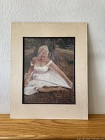 Full view of Marilyn Monroe vintage photograph matted in beige board showing her sitting outside on grass near a tree in a white dress.