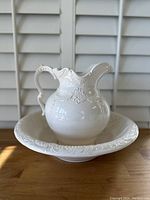 Front view of vintage white ceramic pitcher placed inside matching basin, showing embossed intricate detail and glossy finish.