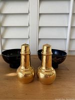 Front view of two brass salt and pepper shakers with two black decorative bowls behind.