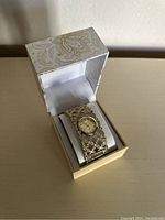 Photo showing the watch in its box from an angled top view, highlighting the ornate gold-tone lattice bracelet and round dial.