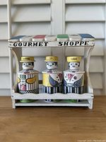 Front view of vintage ceramic coffee, tea and sugar containers with geometric human figure design on white wooden shelf labeled 'Gourmet Shoppe'