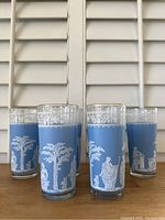 Seven vintage Jeanette Hellenic cobalt blue glasses with white classical Hellenic figures and decorative borders, arranged with three glasses in front and four in back on a wooden surface.