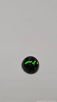 Close up photo of a round black opal gemstone with vibrant flashes of green and hints of red and orange colors, placed on a plain light background.