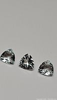 Three natural light-colored, calibrated trillion cut aquamarine gemstones laid out on a neutral gray background showing facets and luster.