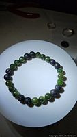 Photo of a natural green jade and black lava stone bead stretchy bracelet laid flat on a white circular surface, showing the alternating pattern and bead details.