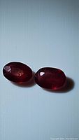 Two oval cut natural rubies on a white background, displaying deep red color, faceting, and clarity enhancements.