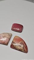 Three polished natural rhodochrosite cabochon stones with varied shapes and patterns on a white surface.