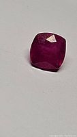 Close up photo of the natural vivid pink-red ruby, showing its cushion shape and faceted polished surface on a plain light background.