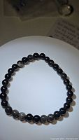 Front view of the round polished black beads with some showing silver sheen reflections on an elastic cord, arranged in a circle.