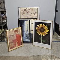 Four framed art prints arranged on floor include sunflower, Whistler print, Eiffel Tower window view, and vintage-style map.