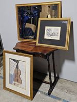 Full view of the lot showing three framed items resting on and near a vintage folding side table against a white wall.