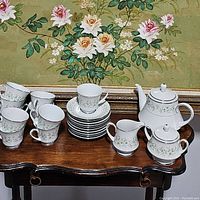 Full tea set displayed on wooden table with floral painting background, includes cups, saucers, creamer, sugar bowl, and teapot