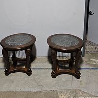Pair of wooden end tables with round removable glass tops showing detailed carvings and metal scrollwork under glass.