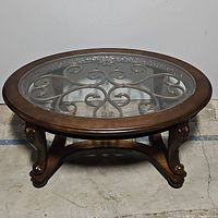 Full view of the oval glass top coffee table showing wood frame, glass surface, and metal scrollwork beneath glass.