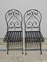 Pair of black iron outdoor chairs with scrollwork backs and lattice seats side-by-side against white background.