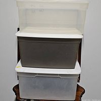 Three plastic storage bins stacked vertically; top clear translucent, middle dark gray/brown opaque, bottom translucent white, all with lids.