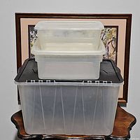 Three stacked clear plastic storage bins on a wooden surface with framed pictures in the background. Smallest bin on top, medium bin in the middle, largest bin on bottom with black lid.