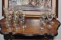 Photo showing two sets of stemware glasses on a wooden table with a framed painting in the background. One set has white floral design and green leaves, the other is plain clear glass.