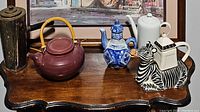 Five assorted tea pots on wooden surface featuring a maroon ceramic pot with bamboo handle, a blue and white porcelain pot, a simple white pot, a tall dark metal or ceramic pot, and a zebra-shaped novelty pot.