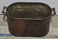 Side angle view of the metal tub showing the oval shape and ornate metal handles on each end, with visible wear and patina on the surface.