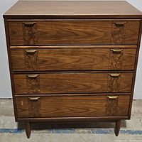 Front view of vintage four drawer wooden chest showing wood grain finish, brass handles, and patterned drawer panels.