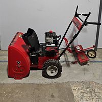 Side view showing red metal body, intake chute, tires, handles, and engine casing.