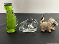 Three cat figurines displayed on a black surface against a white background: green glass cat, clear glass sleeping cat, and small ceramic cats playing.