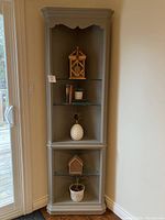 Gray painted corner cabinet with decorative molding, three glass shelves and lighting, shown with decorative items inside but contents not included.