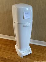 White Diaper Genie diaper disposal system against a beige wall on a wooden floor, with a front view showing the foot pedal and lid.