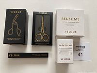 Photo showing Velour branded Too Extra Lash Curler in box, Too Sharp Lash Scissors in box, Reuse Me Cotton Cleansing Pads box, Lash Clean makeup remover box, and Too Easy Lash Applicator box arranged on white surface.