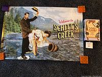 Full view of Schitt's Creek poster pinned at the corners with the Things in a Box game box placed to the right side on floor.