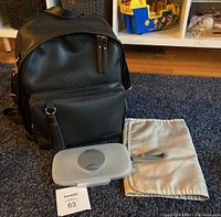 View of black Skip Hop backpack diaper bag with front tassel zipper pocket, gray change pad, gray wipes case with silicone lid, and a beige fabric bag on a dark carpet.
