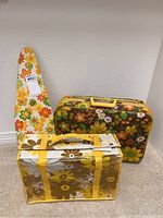 Photo showing three 1980s flower power style items including an ironing board cover, small luggage, and a flat travel bag with yellow straps.